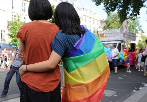 A couple watches a Pride Parade, while wrapped in a rainbow flag (photo credit: Wikimedia Commons)