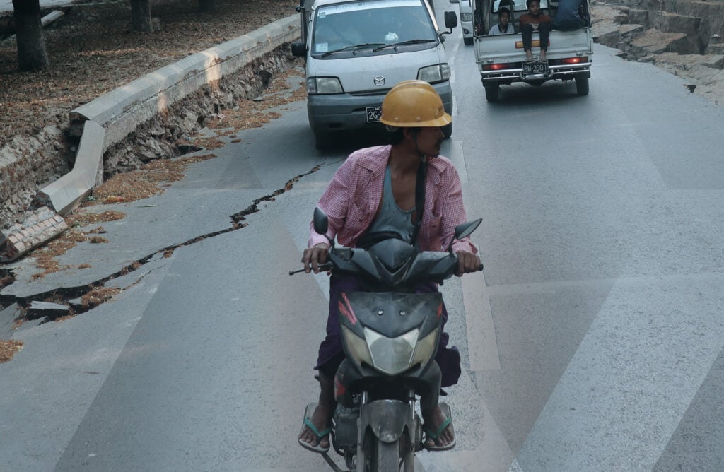 Photo of cars and a man riding a motorbike in the aftermath of the 2025 Myanmar earthquake, with damage visible on the road