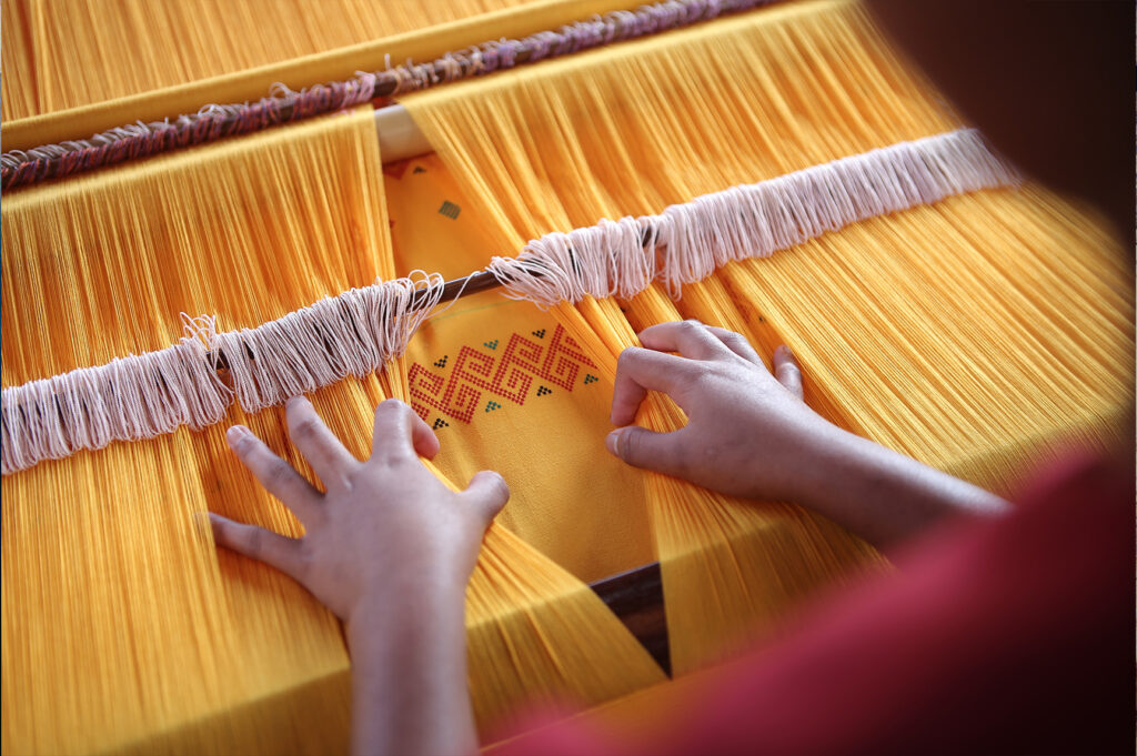 Toraja Indonesia Weaving, showing two hands working a large loom that is strung with mustard yellow thread. (Photo: Aditya Wardhana/Unsplash)