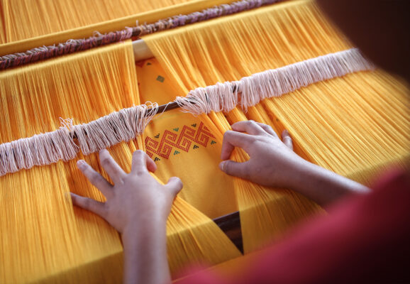 Toraja Indonesia Weaving, showing two hands working a large loom that is strung with mustard yellow thread. (Photo: Aditya Wardhana/Unsplash)