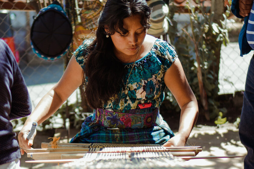 Cultura de Guatemala. A woman with long black hair leans over a large loom, weaving. There are woven items in the background like hats, blankets, and more. Salvadoran CSOs. (Photo: Julio Reynaldo/Unsplash)