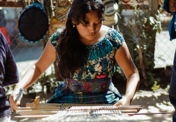 Cultura de Guatemala. A woman with long black hair leans over a large loom, weaving. There are woven items in the background like hats, blankets, and more. (Photo: Julio Reynaldo/Unsplash)
