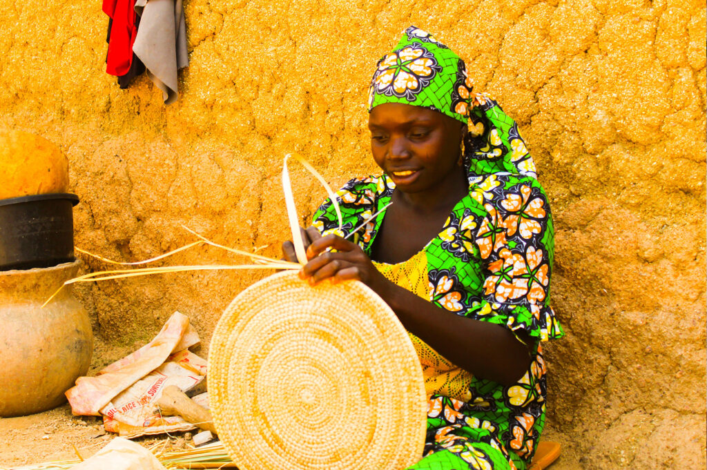 A young woman wearing bright green weaves a natural fiber basket in Kano, Nigeria in front of a bright yellow wall. Nigerian legal reform. (Photo: Nnaemeka Ugochukwu/Unsplash)