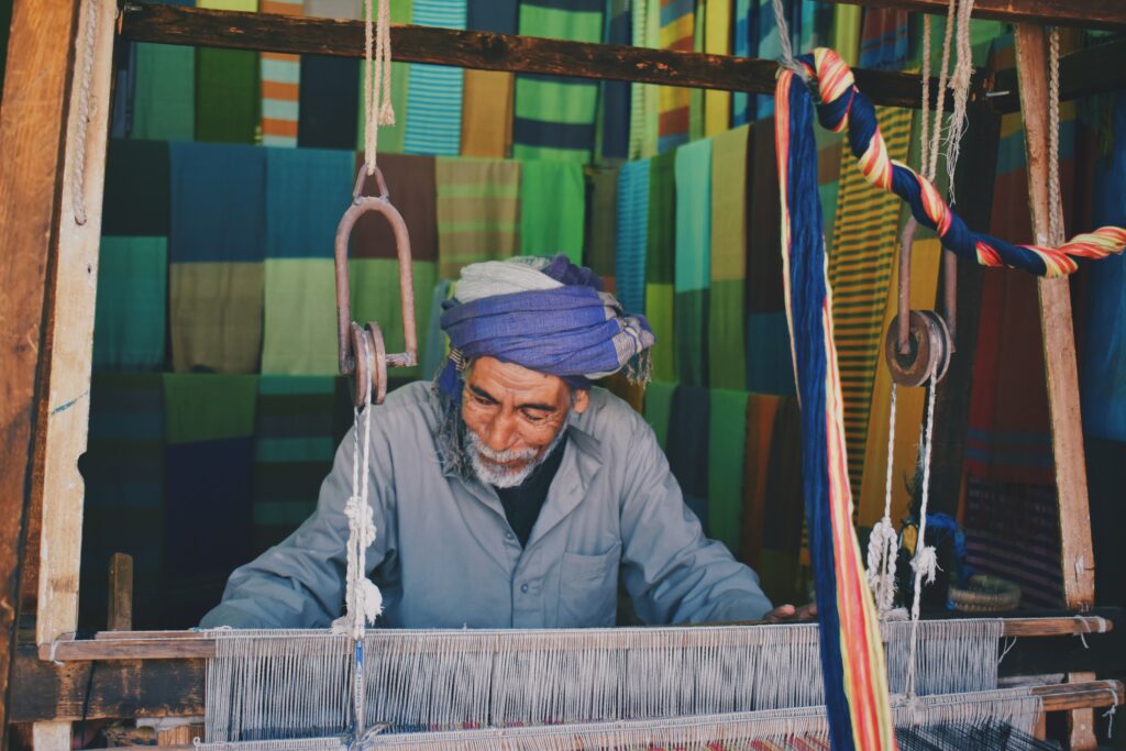 Man wearing a blue turban is weaving rugs on a large traditional loom in Aswan, Egypt; safeguarding assembly rights worldwide. (Photo: Rawan Yasser/Unsplash)