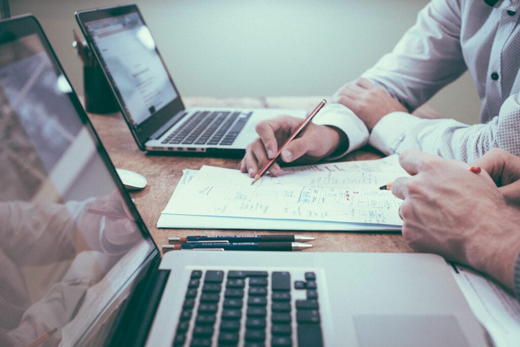 Two people sitting at a table with open laptops. They are taking notes on a notepad between the laptops. Only their hands are visible. FATF Recommendation 8.